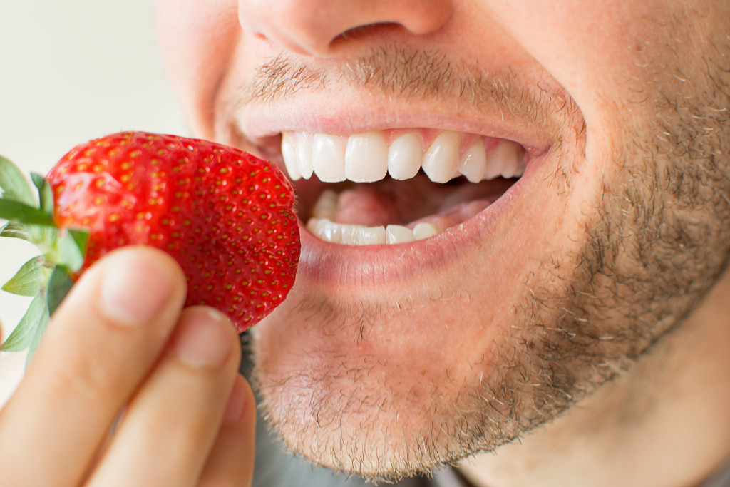 Strawberry near the mouth of a young man who's ready to eat it.