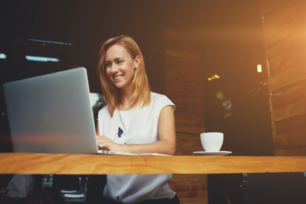 A freelance writer smiling while typing in a cafe with a cup of coffee beside her