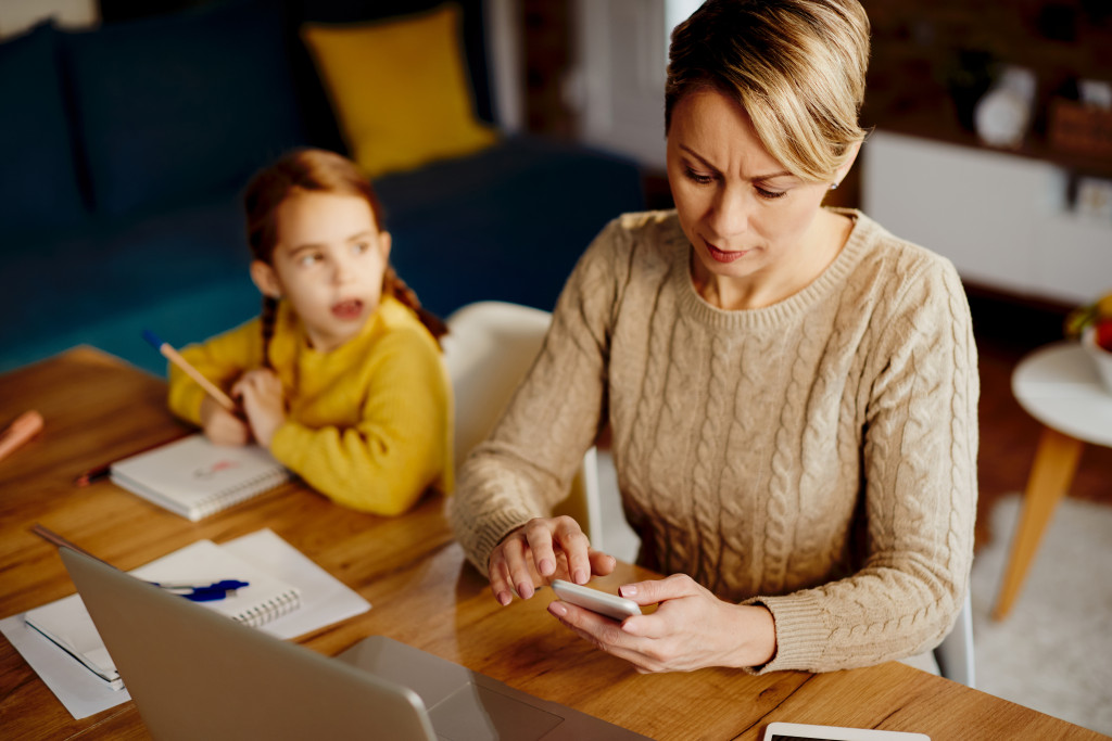 Young mother checking messages on her phone while working from home with her daughter doing homework beside her.