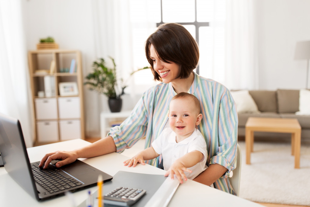 a mother using a laptop while holding her kid at home