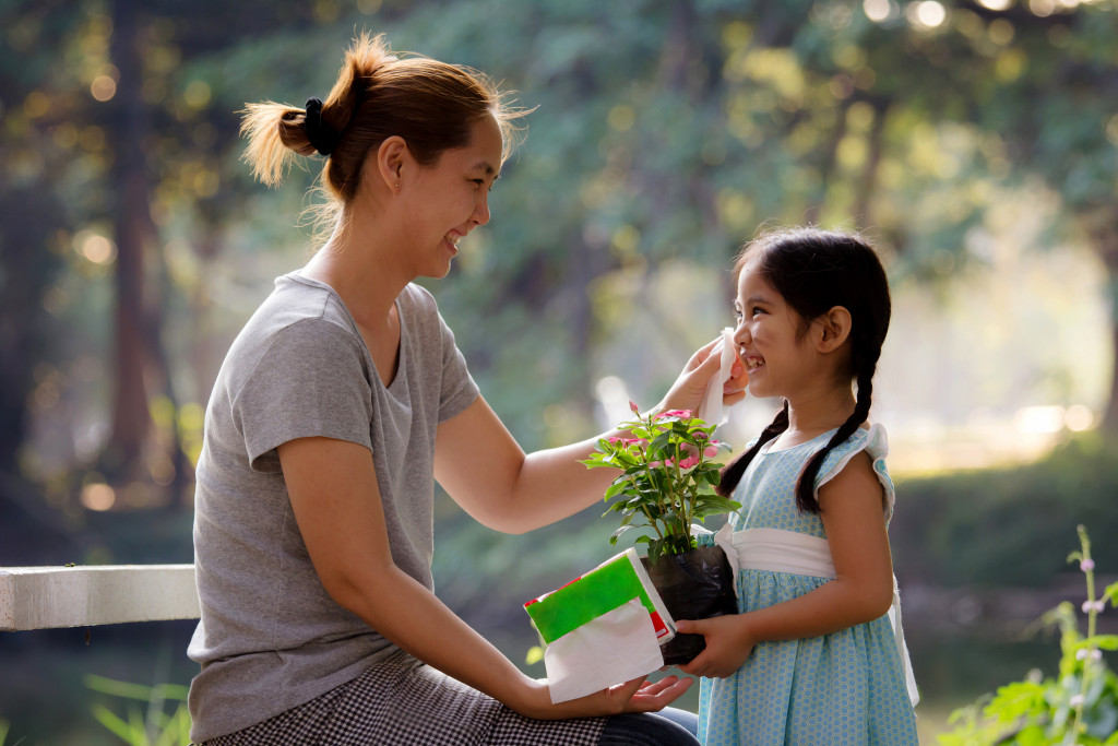 A mother wipes dirt off her daughter's cheek using a tissue while the little girl holds a potted plant in her hands