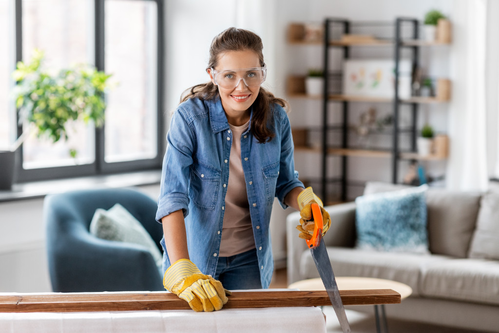 A woman sawing wood at home while wearing gloves and protective goggles