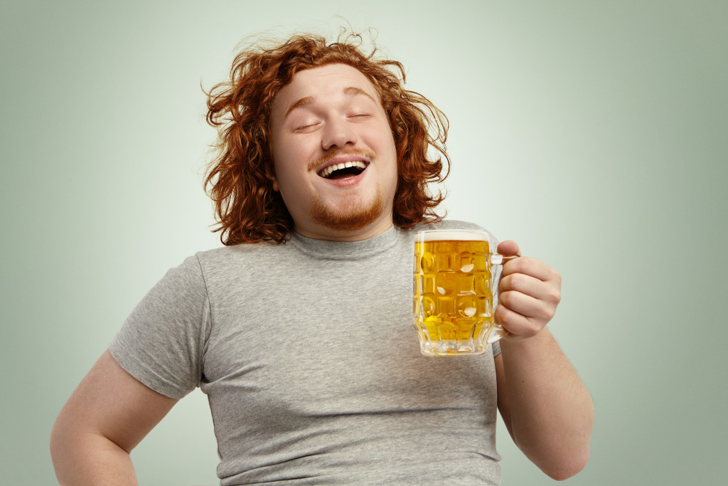 Funny young Caucasian male feeling happy and relaxed, anticipating fresh cold beer in his hands after hard working day, closing eyes in enjoyment