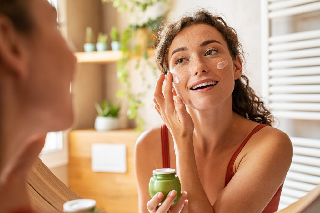 a woman in a bathroom looking at the mirror while applying a cream from a bottle