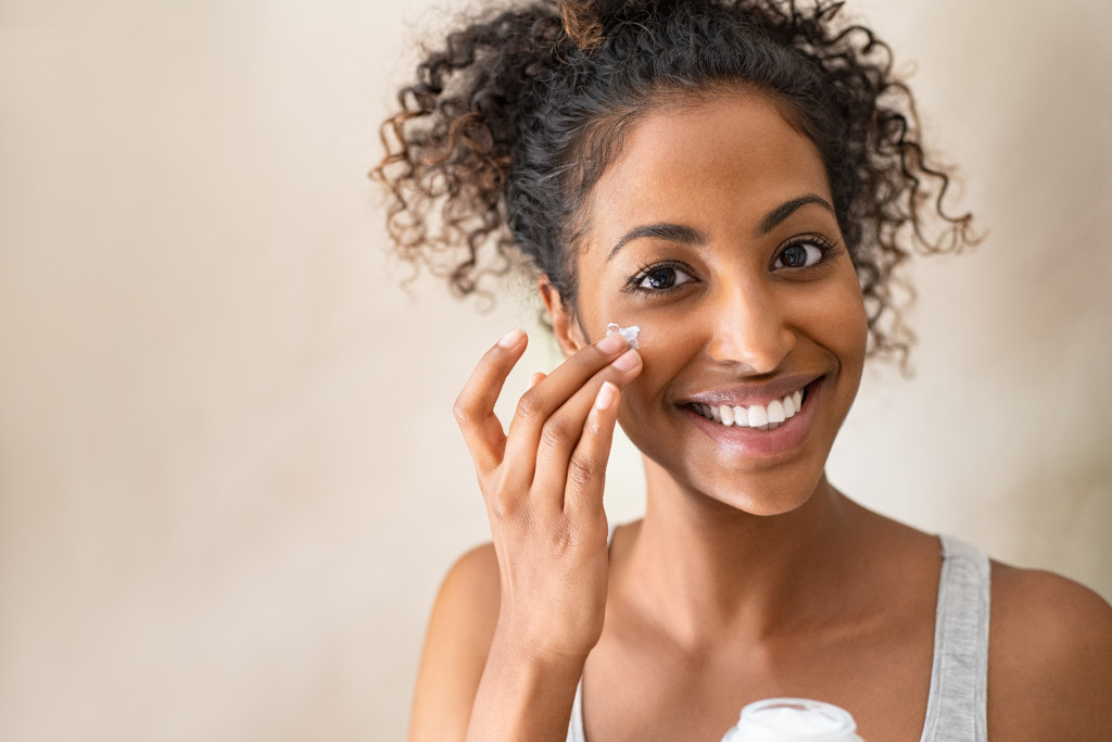 Portrait of a young black woman applying cream on her face isolated on beige background