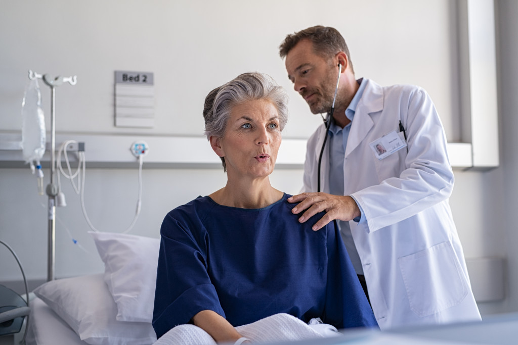 Hospitalized elderly woman sitting on bed while doctor checking her breathe and heartbeat