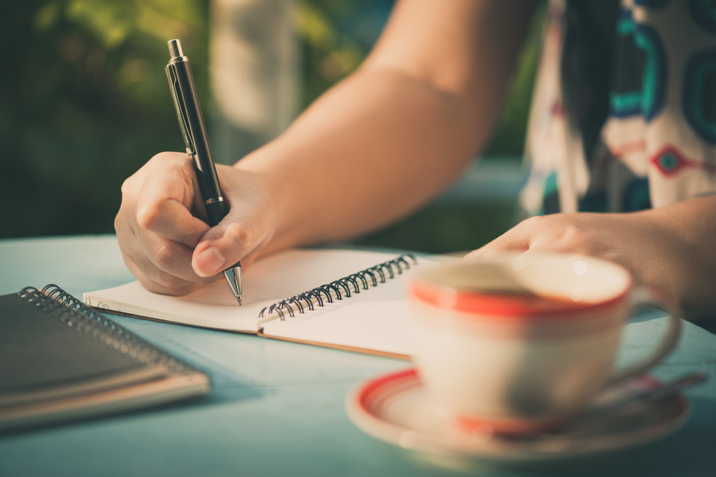 A woman writing on a notebook while drinking coffee